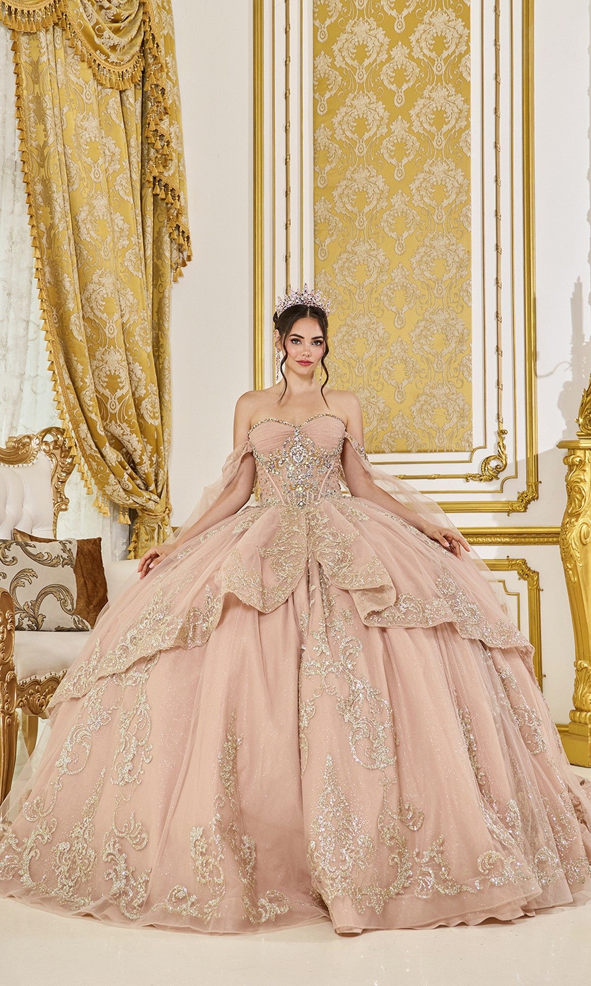 A woman stands in an ornate room wearing the rose gold Quinceanera Dress 5069 by Dancing Queen, featuring off-the-shoulder sparkling embellishments and a tiara.